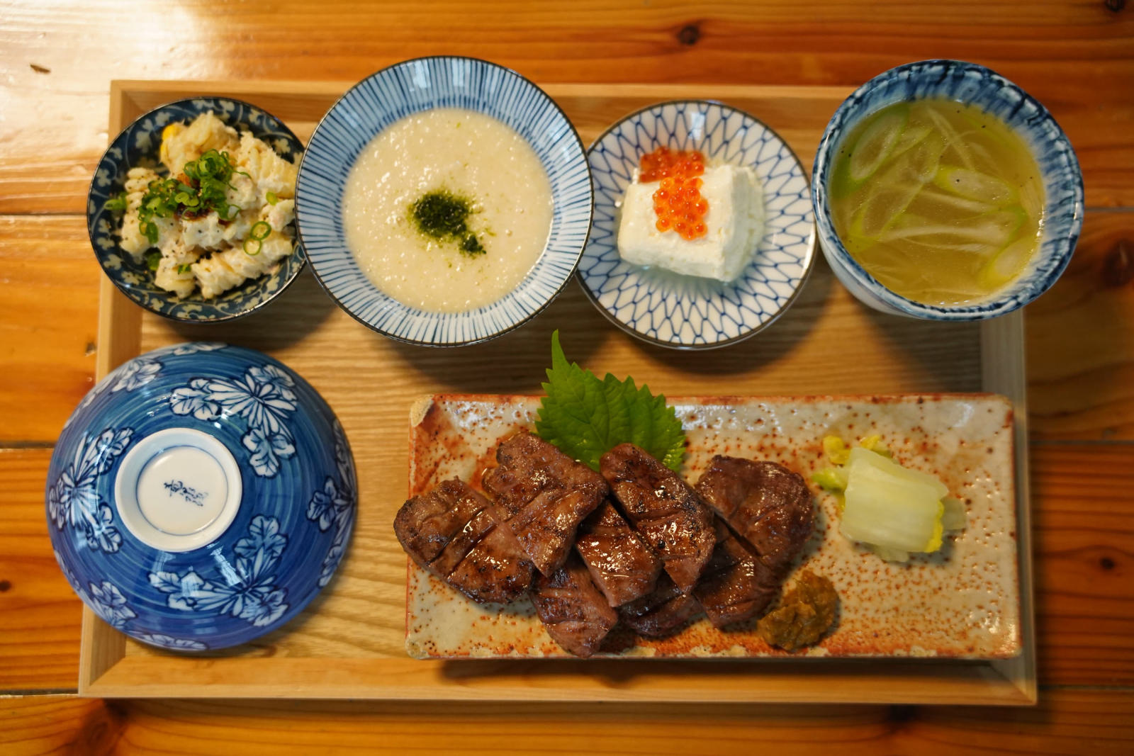 牛たん定食の膳を真上から。大葉を添えた牛たん焼き、白菜の漬物と南蛮味噌、とろろ、いくらをのせた豆腐、スープ、小鉢、伏せた染付の飯碗。