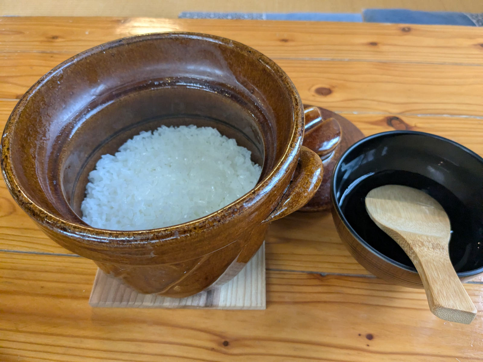 木の敷板にのせた土鍋の白いご飯。隣に黒い椀としゃもじ。