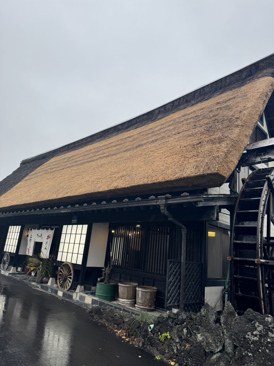 曇り空の下、長く伸びる茅葺き屋根。入口に「たん平」「牛たん」の暖簾、障子窓と格子の外壁、壁沿いに荷車の車輪と木桶、右に水車、濡れた路面。