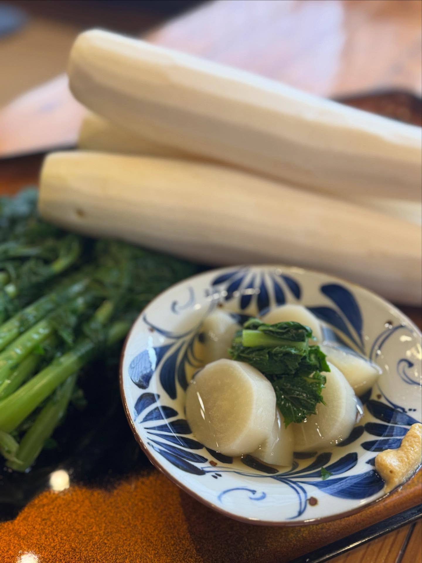 藍の絵付けの小鉢に大根と葉の煮物。奥に皮をむいた大根と青菜。